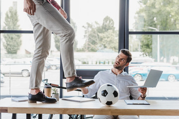 cropped shot of man kicking soccer ball on table while colleague using laptop in office