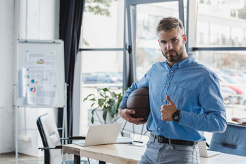 handsome young businessman holding basketball ball and showing thumb up in office