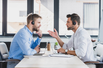 side view of young call center operators in headsets discussing and looking at each other while using laptops in office