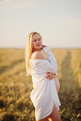 Fototapeta na wymiar Blonde pregnant woman in wreath field at white underwear clothes on sunset. Happy moments of pregnancy.