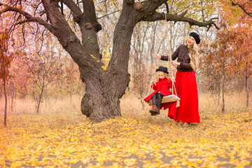 beautiful mother in a long dress is swinging on a hinged swing little daughter in a red coat in an autumn garden near an old gnarled tree