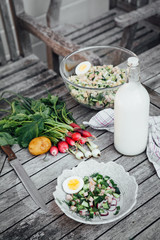 A bottle of fermented milk, sliced vegetables and sausages, ingredients for a cold summer soup. On a wooden table in the garden