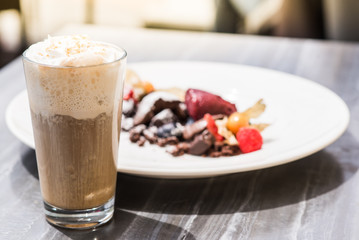 Vienna coffee - hot coffee with whipped cream on blurred background of chocolate cake with forest fruit on a white dessert plate in an outdoor cafe.