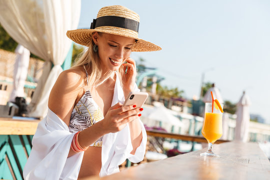 Photo of gorgeous woman 20s in straw hat smiling and holding smartphone, while drinking orange juice in beach bar during summer sunny day