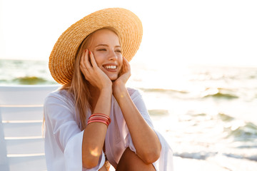 Portrait of european cheerful woman 20s in straw hat smiling, while sitting in lounge chair at seaside during summer morning