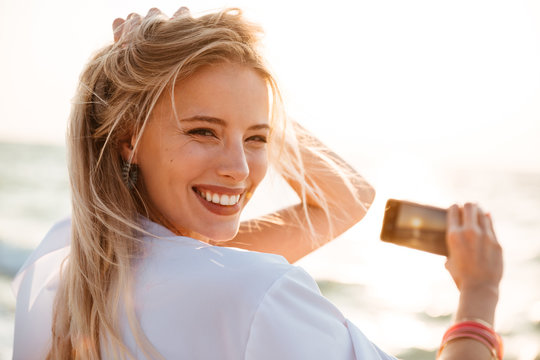 Photo of joyous blonde woman 20s smiling, and taking photo of sunrise above sea, while walking on summer beach