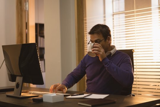 Businessman having coffee while using desktop pc at desk