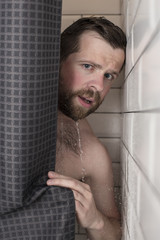 Handsome, bearded man with wet hair looks from behind a curtain in the shower. Close-up.