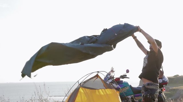 Dude, the biker puts up a tent, a strong wind blows. He's got a mustache, a beard, and a black baseball cap on his head. He stands near his motorcycle enduro on a cliff against the sea.