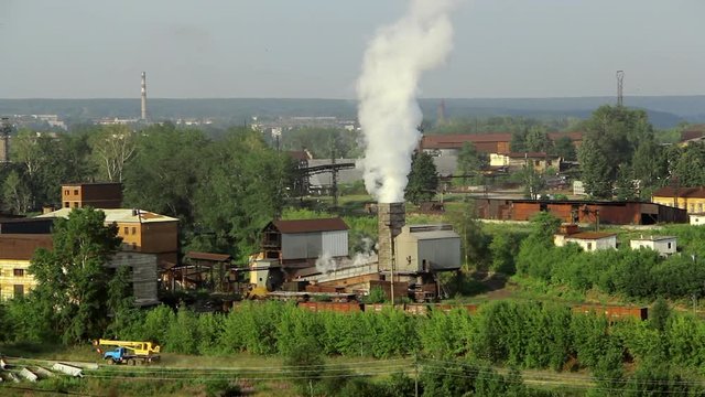 Pipes of industrial plant smoke among green trees and nature in Alapaevsk. Industrial zone in remote corner of Russia. Alapaevsky Metallurgical Plant pollutes environment of small city of Ural.