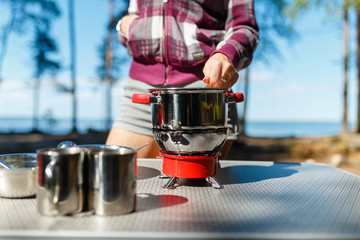 Girl traveler prepares food on portable gas stove, on a folding table on the background of camping in forest. Women's hands interfere with a spoon food in a pot on a gas burner (camping stove).