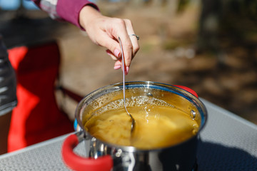 Girl traveler prepares food on portable gas stove, on a folding table on the background of camping in forest. Women's hands interfere with a spoon food in a pot on a gas burner (camping stove).