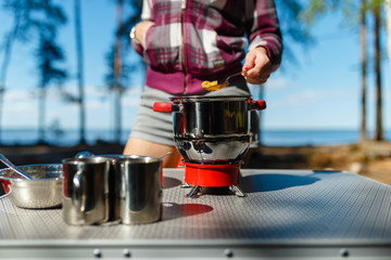 Girl traveler prepares food on portable gas stove, on a folding table on the background of camping...