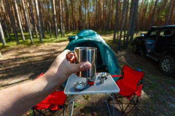 The man's hand holds an iron mug with a warm drink on the background of the table and chairs, campfire, a tent and an SUV in camping. Family vacation, concept of traveling by SUV or car in the wild.