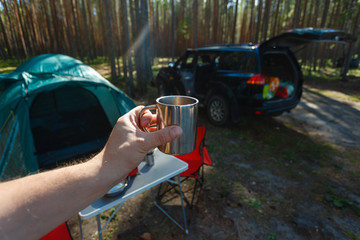 The man's hand holds an iron mug with a warm drink on the background of the table and chairs, campfire, a tent and an SUV in camping. Family vacation, concept of traveling by SUV or car in the wild.