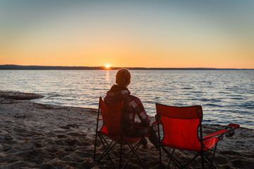 Girl traveler on a folding chair admiring the sunset on the lake. Young woman sitting alone in a folding chair watching the sun setting on the shore at the camping.