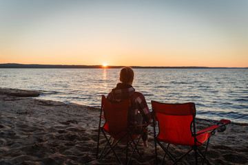 Fototapeta premium Girl traveler on a folding chair admiring the sunset on the lake. Young woman sitting alone in a folding chair watching the sun setting on the shore at the camping.