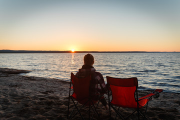 Girl traveler on a folding chair admiring the sunset on the lake. Young woman sitting alone in a folding chair watching the sun setting on the shore at the camping.