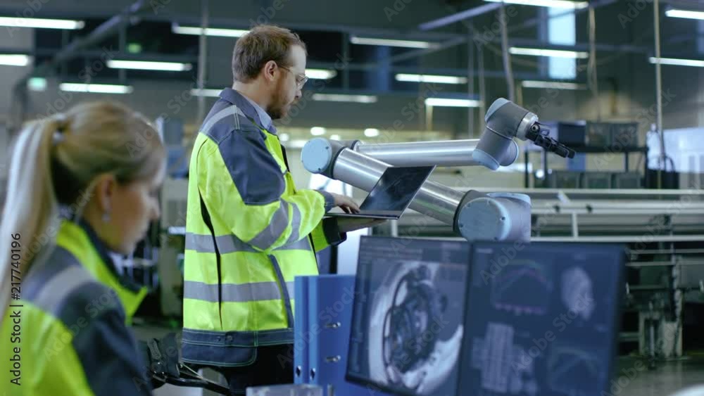 Poster At the Factory: Male Automation Engineer Uses Laptop for Programming Robotic Arm while Female Mechanical Engineer Designs 3D Engine on Her Personal Computer.