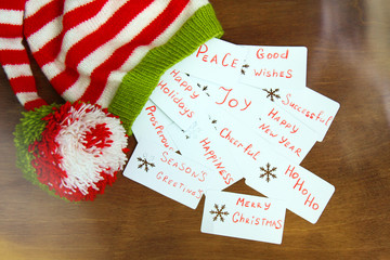 Fototapeta premium Colorful christmas red, white and green knitted Santa claus helper hat, natural wool, handmade knitted, with christmas wishes on paper cards with snowflakes on brown wooden background.
