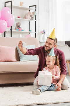 father and adorable daughter in party hats taking selfie with gift for birthday