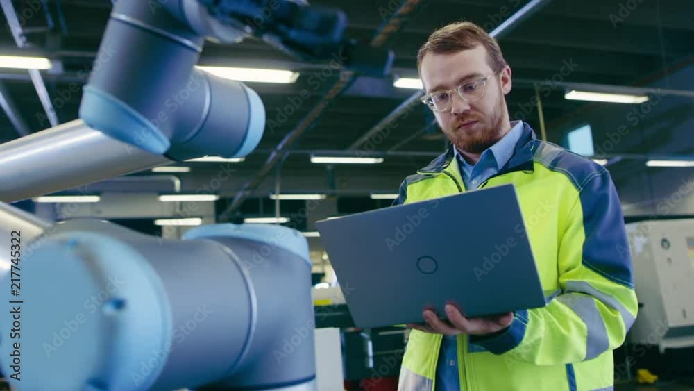 Poster Low Angle Shot At the Factory: Automation Engineer Uses Laptop for Programming and Testing Robotic Arm. New Era in Automatic Manufacturing Industry.