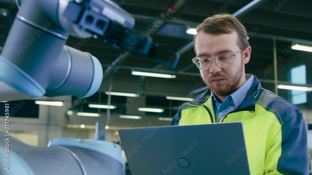 Poster Handheld Shot At the Factory: Automation Engineer Uses Laptop for Programming and Testing Robotic Arm. New Era in Automatic Manufacturing Industry.