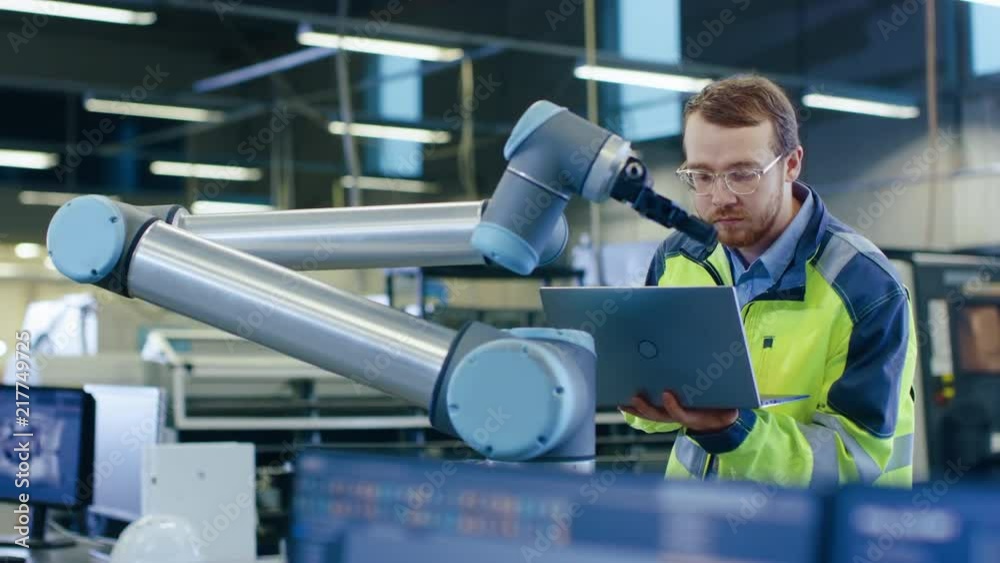 Poster At the Factory: Automation Engineer Uses Laptop for Programming Robotic Arm. New Era in Automatic Manufacturing Industry. Shot on RED EPIC-W 8K Helium Cinema Camera.