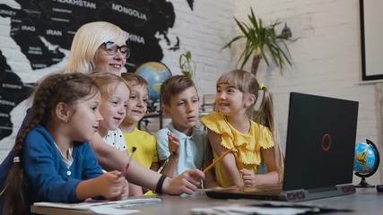 Teacher sitting at desk next to students as they discuss information on laptop. Pupils with teacher are using the black laptop together in the geography classroom in elementary school