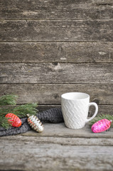 White cup with pattern, Christmas tree decoration cones and green pine branches on a knitted sweater. Wooden gray background.