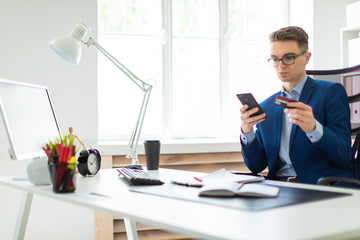 A young man sits at a table in the office and holds a bank card and phone.