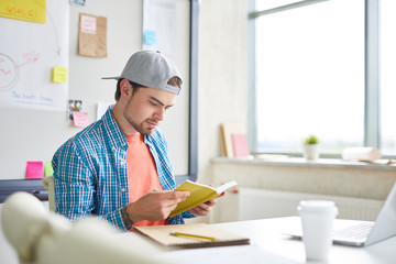 Young man in casualwear sitting by desk and reading book while preparing for lesson in classroom