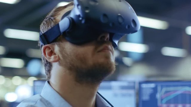 Close-up Portrait of the Industrial Engineer Putting on Virtual Reality Headset, ready to Work. In the Background Manufacturing Plant and Monitors.