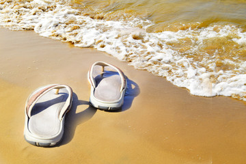 Slippers are on wet sand near a water edge. Flip-flops are on the beach. Wave with white foam of the Baltic Sea.