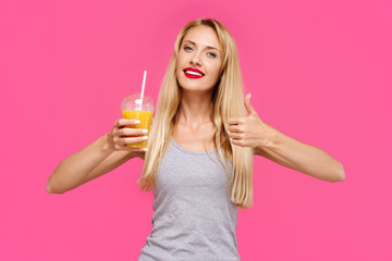 Young beautiful slender girl with a pink background holding orange juice smiling and showing thumb up. Concept of un healthy fat junk and healthy food