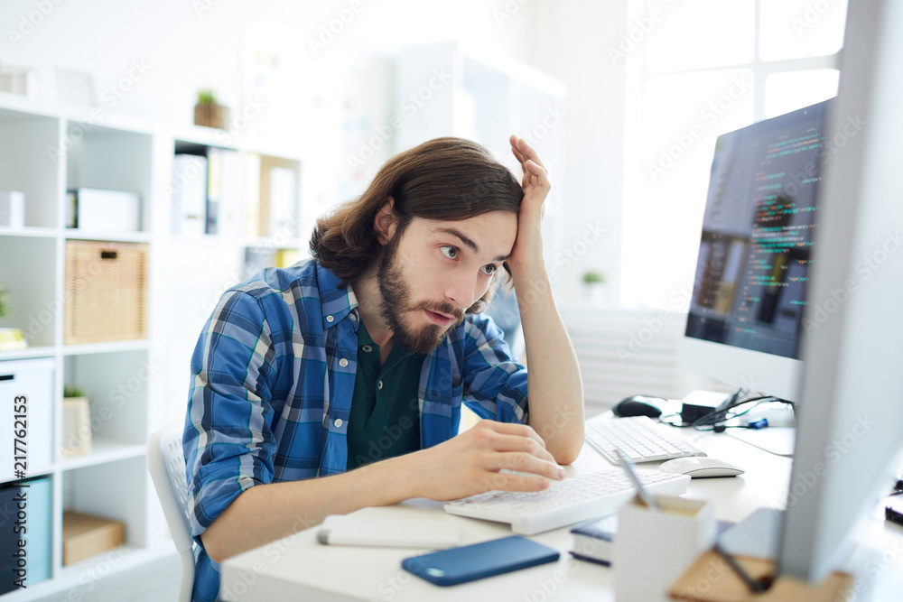 Wall mural young confused it-manager looking at one of computer monitors with tense expression