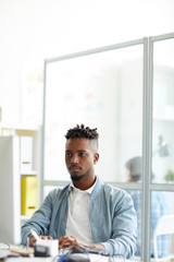 Young African-american hipster sitting by workplace in front of computer monitor and learning new...