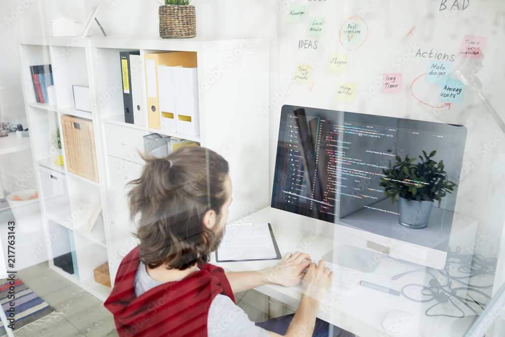 Wall mural young it-manager sitting in front of monitor, typing and looking at coded data on screen