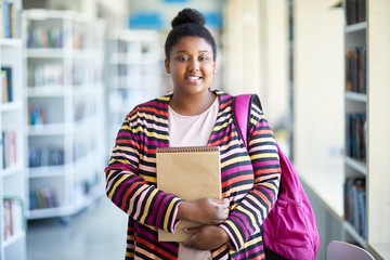 Positive confident overweight African-American student girl in colorful cardigan holding sketchpad and smiling at camera in library