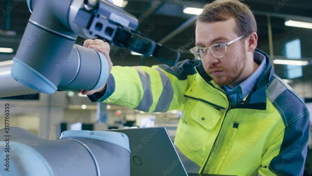 Leinwandbilder At the Factory: Automation Engineer Uses Laptop for Programming Robotic Arm. New Era in Automatic Manufacturing Industry.