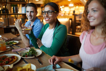 Pretty young woman with drink sitting by served table among her friends and listening to one of them