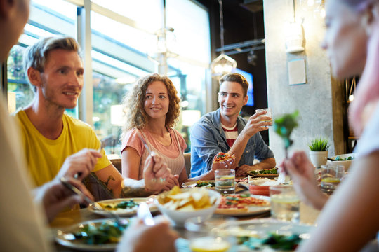Cheerful young friendly people having talk by table served with tasty meal in cafe or restaurant
