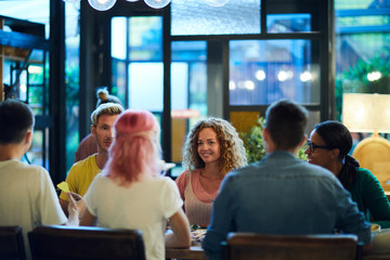 Happy intercultural young friends gathered by table in cafe for talk and dinner