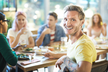 Happy guy with toothy smile turning for camera while sitting by sersved table among his friends