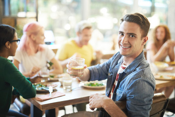 Young smiling man cheering up with drink while looking at you during dinner with friends