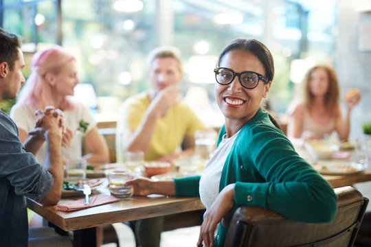 Mixed-race girl with toothy smile looking at you while sitting by served table at dinner