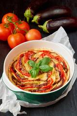 Traditional French dish quiche - pie with eggplants, tomatoes and cheese in the green baking dish decorated with fresh basil leaves. Homemade vegetable tart on the black background. Close up view