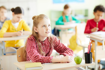 Pensive cute schoolgirl in casual shirt attentively listening to teacher while sitting at desk with school supplies and apple at lesson