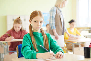 Serious confident schoolgirl with red hair sitting at table and holding pencil while passing exam in classroom, she looking at camera