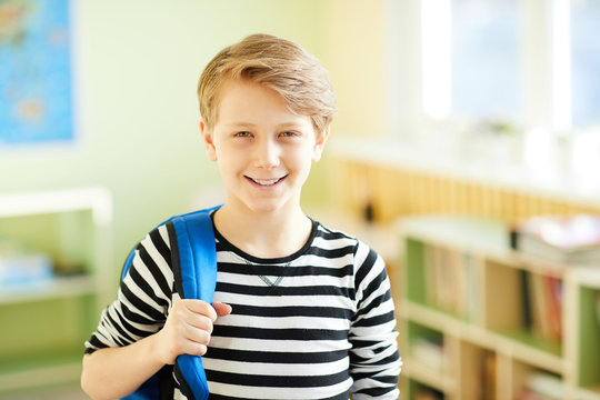Cheerful confident male pupil holding handle of satchel and looking at camera while standing in school corridor